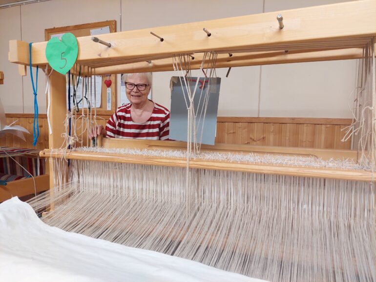 vanhempi nainen kangaspuiden takana kutomassa.