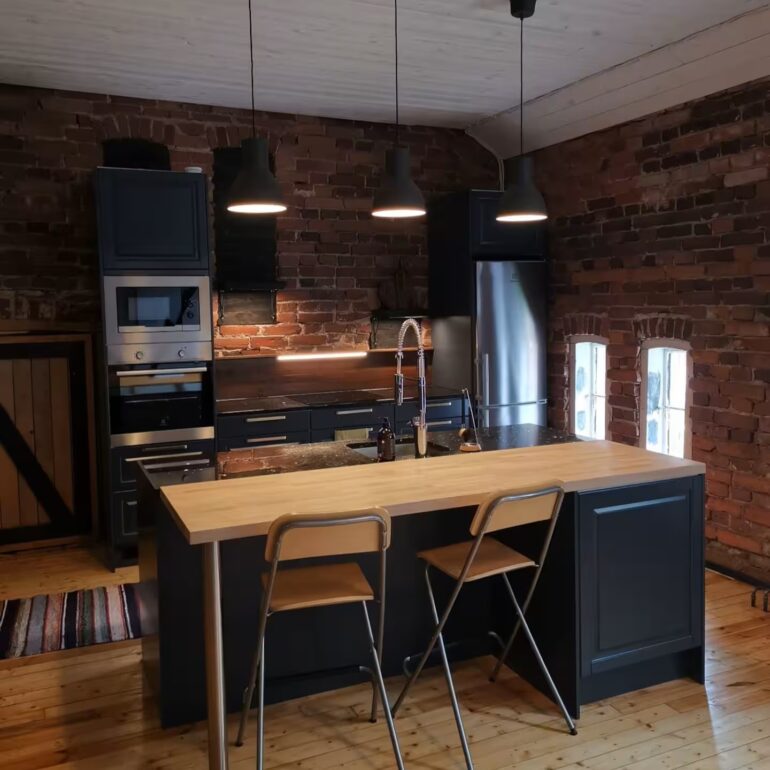 Musta, moderni keittiö punaista tiiliseinää vasten. Keittiön saarekkeen edessä kaksi tuolia.