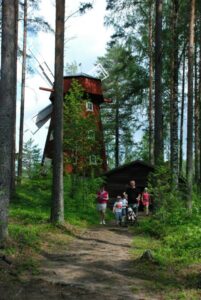 Museovieraita, lapsia ja aikuisia kävelee Laurinmäellä taustallaan sepän hirsinen paja ja punainen tuulimylly.