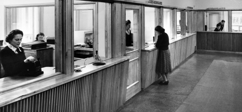 Konttorin aulasta otettu kuva vuodelta 1949. Kassaluukulla työskentelee nainen kassakoneen ääressä, huoltoluukulla asioi toinen nainen.