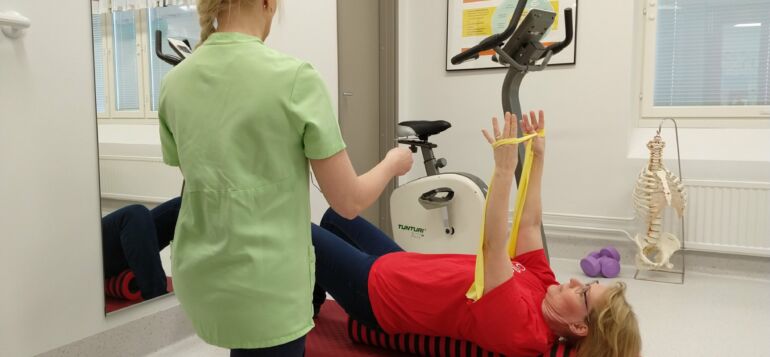 Fysioterapeutti ohjaa asiakkaalle kuminauhaharjoitusta valoisassa terapiatilassa.