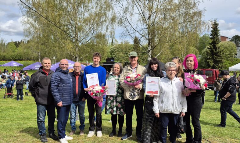 Ihmisiä rivissä nurmialueella keväisessä auringonpaisteessa. Taustalla muita ihmisiä ja kaksi isoa koivua.