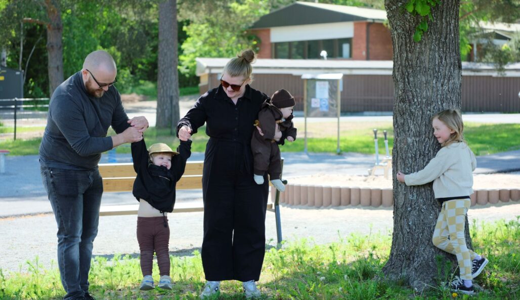 A mom and a dad lifting up a child by his arms. Mom is also carrying a baby in her arm. A little older child is hugging a tree next to the parents.