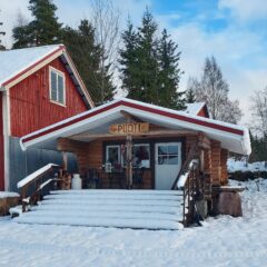 Pieni puinen aitta talvisessa maisemassa. Vieressä kohoaa suurempi punainen talo. Aitan seinässä teksti Puoti.