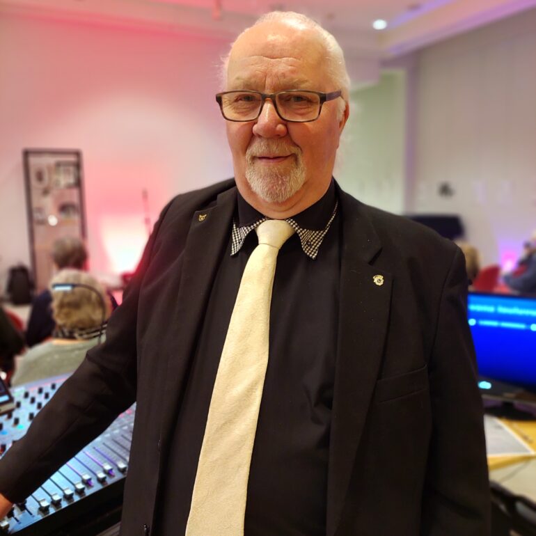 Silmälasipäinen vanhempi mies mustassa puvussa.