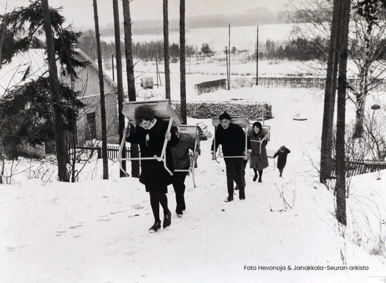 Mustavalkoinen valokuva, jossa etualalla kolme oppilasta kantaa selässään pulpettejaan ylös mäkeä. On talvi ja lunta on maassa. Oppilaat hymyilevät.