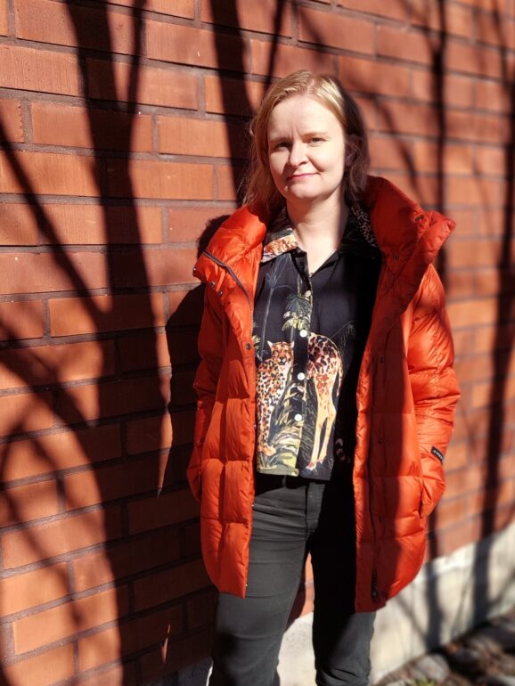 Henkilökuva hallinto- ja henkilöstöjohtaja Maija Nivalasta. Nivala seisoo puiden varjossa punatiilisen rakennuksen edessä, yllään oranssi takki ja mustat housut.