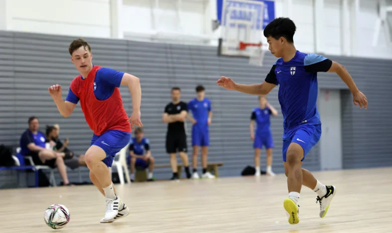 Two futsal players reaching for a ball. Other players watch in the background.