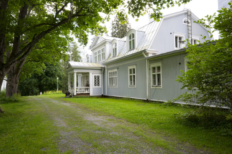 Vaaleansininen puinen kartano ja sen etupiha. Kaksi isoa puuta kartanon edessä, jotka tekevät varjoisen sisääntulon taloon.