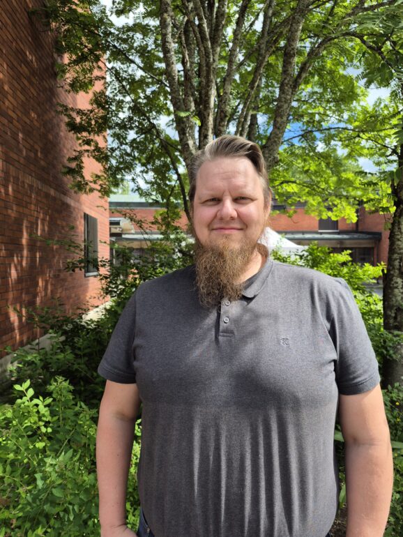 Puolilähikuva miehestä, jolla on harmaa paita ja vaaleanruskea parta. Taustalla punaista tiiliseinää ja vehreää puustoa ja pensaikkoa.