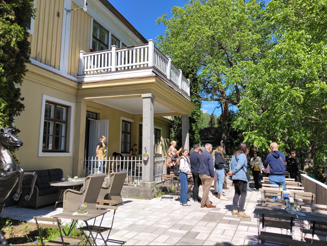 Ihmisiä seisoo suuren keltaisen rakennuksen laatoitetulla pihalla sen edustalla.