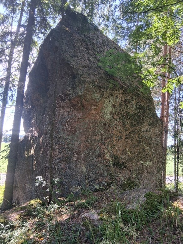Korkea kivi kohoaa puiden lomassa. Kiven takana näkyy peltomaisema.